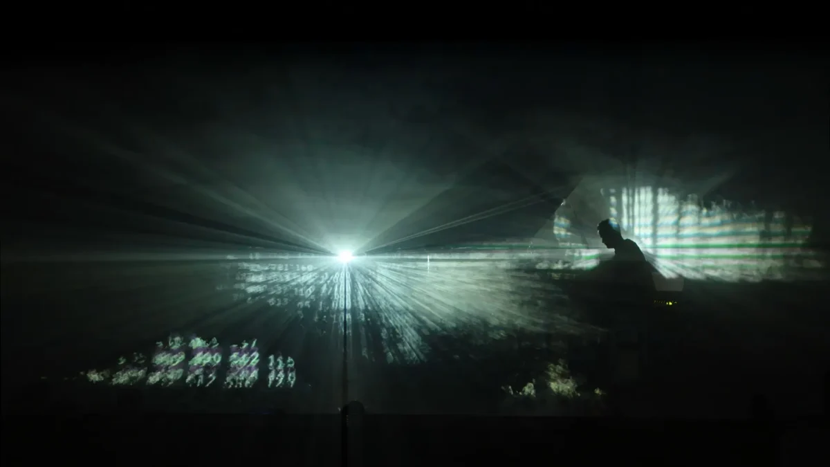 Projected light rays and silhouette during "Da Lontanissimo," a sequence within Per Aria
Image Caption: "Da Lontanissimo" — light and data-like projections unfold across the performance space.